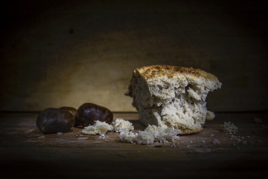 A single chunk of bread lies on a worn wooden table next to some chestnuts in a dark and moody room. The bread has been picked up so there are a few crumbs around. Was this from a midnight snack? Or could this be an offering for unknown spirits.