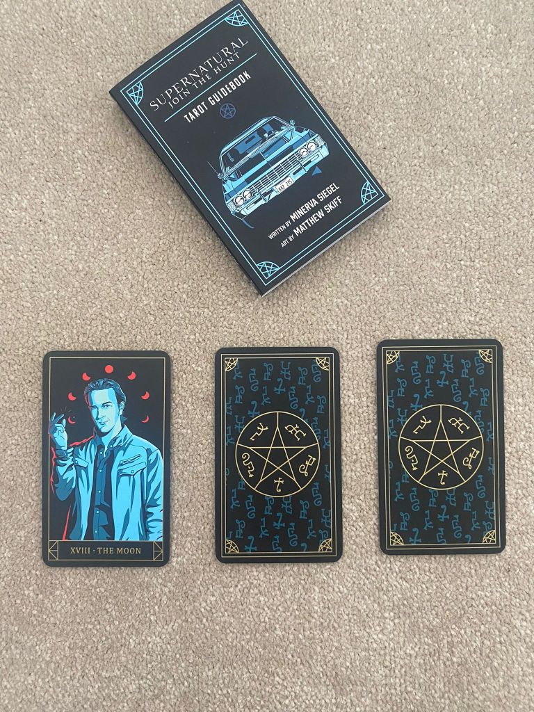 The Supernatural Tarot deck and guide book is spread out on the carpet. the first card is turned up showing XVIII The Moon - represented here by an illustrated version of the character Loki / Gabriel in various shades of blue with the red phases of the moon circling his head. 2 cards remain face down with a pentacle in the centre.