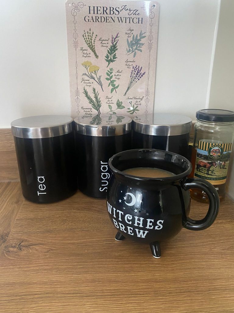 A black coffee cup that looks like a cauldron holds a cup of tea and says 'witches brew' on the front